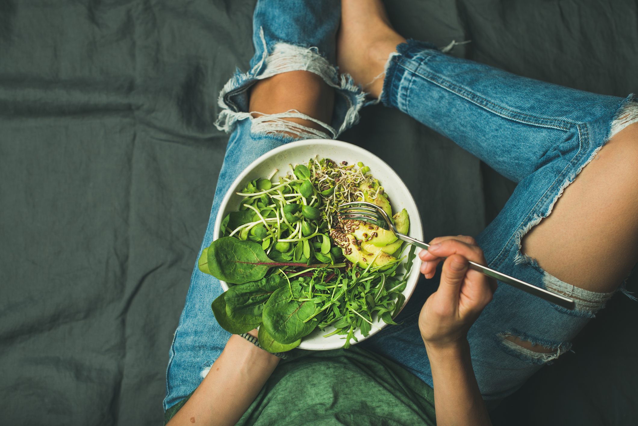 Green vegan breakfast meal in bowl with spinach, arugula, avocado, seeds and sprouts. Girl in jeans holding fork with knees and hands visible, top view, copy space. Clean eating, vegan food concept