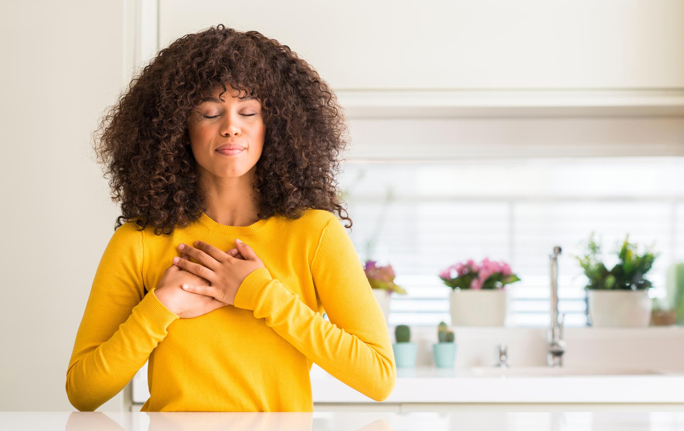African american woman wearing yellow sweater at kitchen smiling with hands on chest with closed eyes and grateful gesture on face. Health concept.